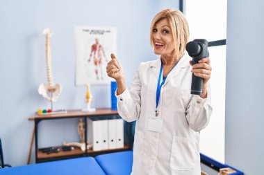 Middle age blonde woman holding therapy massage gun at physiotherapy center smiling happy and positive, thumb up doing excellent and approval sign 