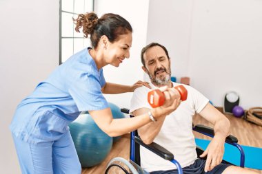 Orta yaşlı bir adam ve kadın, fizyoterapi kliniğinde tekerlekli sandalyede oturan dambılı kullanarak rehabilitasyon seansı yapıyorlar.