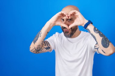 Mavi arka planda dikilen İspanyol dövmeli adam. El ve parmaklarıyla kalp şekli yapıyor. İşaretten bakıp gülümsüyor. 