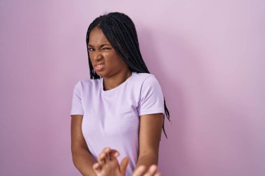African american woman with braids standing over pink background disgusted expression, displeased and fearful doing disgust face because aversion reaction. 