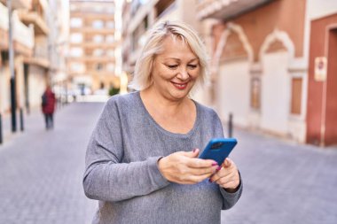 Orta yaşlı sarışın kadın sokakta akıllı telefon kullanarak gülümsüyor.