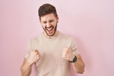 Pembe arka planda duran İspanyol sakallı adam mutlu ve heyecanlı bir şekilde kolları havada, gülümseyen ve başarı için bağıran kazanan bir jest yapıyor. kutlama konsepti. 