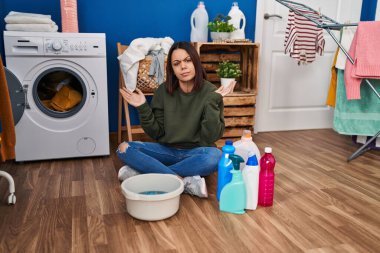 Young hispanic woman doing laundry washing by hand clueless and confused expression with arms and hands raised. doubt concept. 