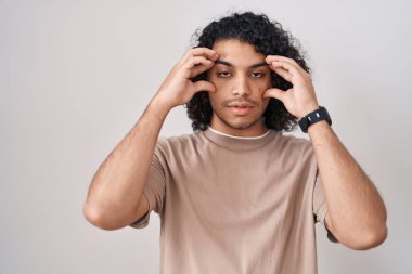 Beyaz arka planda duran kıvırcık saçlı İspanyol adam parmaklarıyla gözlerini açmaya çalışıyor, uykulu ve yorgun. 