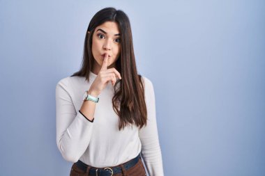 Genç esmer kadın mavi arka planda dikiliyor ve parmakları dudaklarında sessiz olmak istiyor. sessizlik ve gizli konsept. 