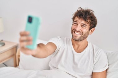 Genç adam yatakta akıllı telefonuyla selfie çekiyor.