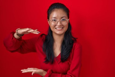 Asian young woman standing over red background gesturing with hands showing big and large size sign, measure symbol. smiling looking at the camera. measuring concept. 