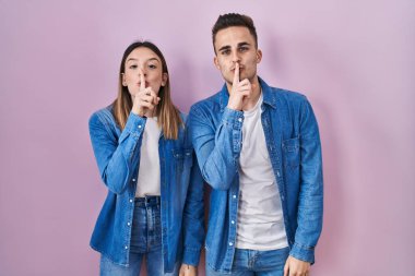 Young hispanic couple standing over pink background asking to be quiet with finger on lips. silence and secret concept. 