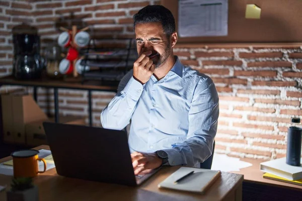 Hispanic man with beard working at the office at night smelling something stinky and disgusting, intolerable smell, holding breath with fingers on nose. bad smell