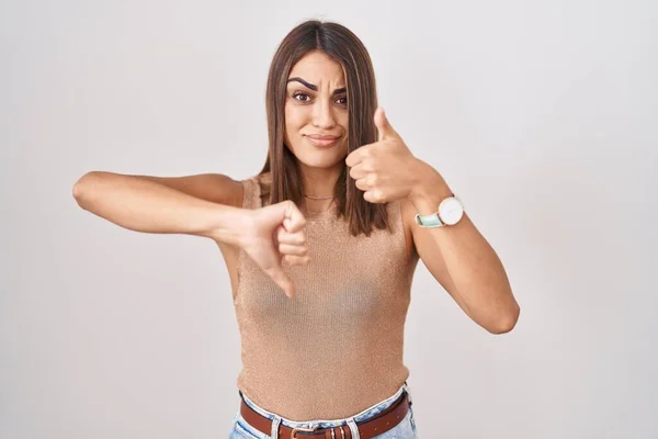 Young hispanic woman standing over white background doing thumbs up and down, disagreement and agreement expression. crazy conflict