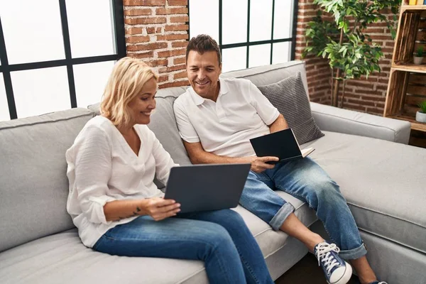 Middle age man and woman using laptop and reading book sitting on sofa at home