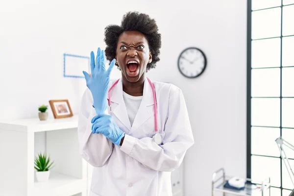 African doctor woman wearing latex gloves at medical clinic angry and mad screaming frustrated and furious, shouting with anger. rage and aggressive concept.