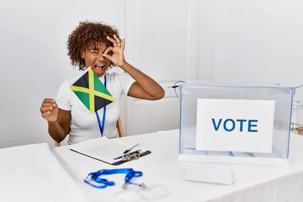 Young african american woman at political campaign election holding jamaica flag smiling happy doing ok sign with hand on eye looking through fingers