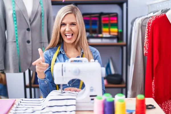 Blonde woman dressmaker designer using sew machine pointing fingers to camera with happy and funny face. good energy and vibes.