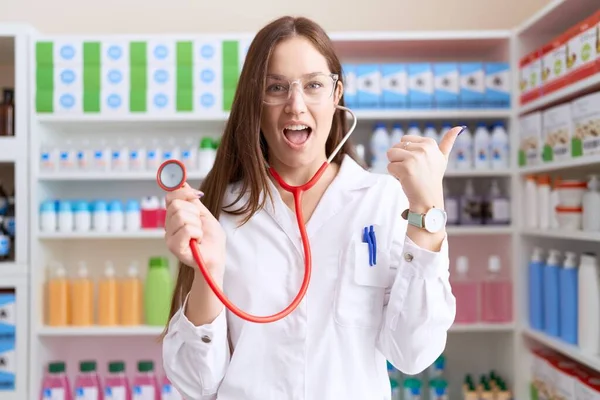 美しいですブルネット女性仕事で薬局ドラッグストア保持Stethoscope指して親指へザサイド笑顔幸せとともにオープン口 — ストック写真