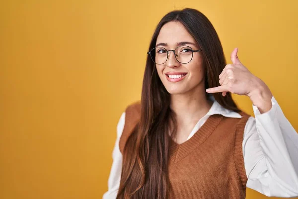 年轻的黑发女人站在黄色的背景上 戴着眼镜 微笑着 用手和手指做着电话手势 就像在电话里说话一样 交流概念 — 图库照片