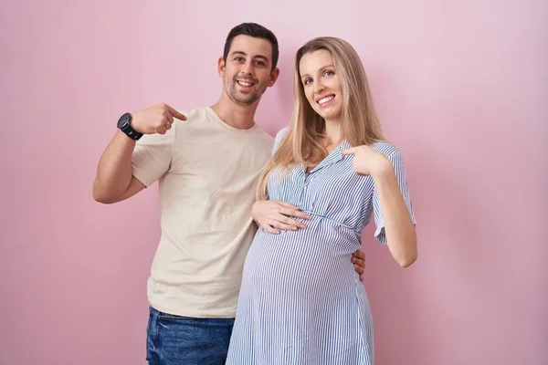 顔に笑顔で自信を持っているように見えるピンクの背景に立っている赤ちゃんを期待若いカップル 誇りと幸せ指で自分自身を指して — ストック写真
