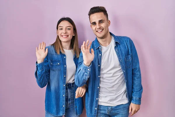 Young Hispanic Couple Standing Pink Background Waiving Saying Hello Happy — Stockfoto