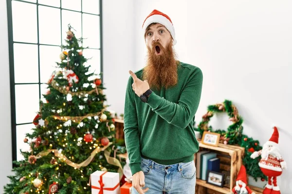 赤毛男とともに長いひげ身に着けているクリスマスの帽子によってクリスマスツリー驚くべきポインティングとともに指で側面 口を開けて驚くような式 — ストック写真