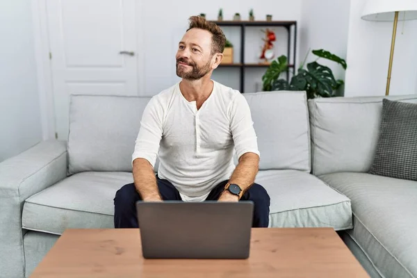 Middle age man using laptop at home smiling looking to the side and staring away thinking.