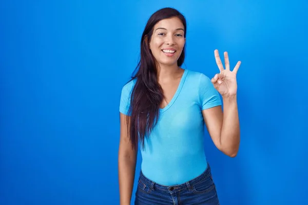 若いですヒスパニック女性立ちます上の青の背景表示と指番号3を指しますしながら笑顔自信と幸せ — ストック写真