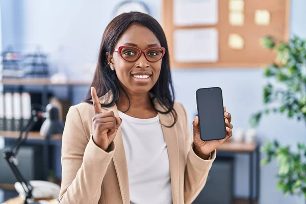 African young woman holding smartphone showing blank screen surprised with an idea or question pointing finger with happy face, number one
