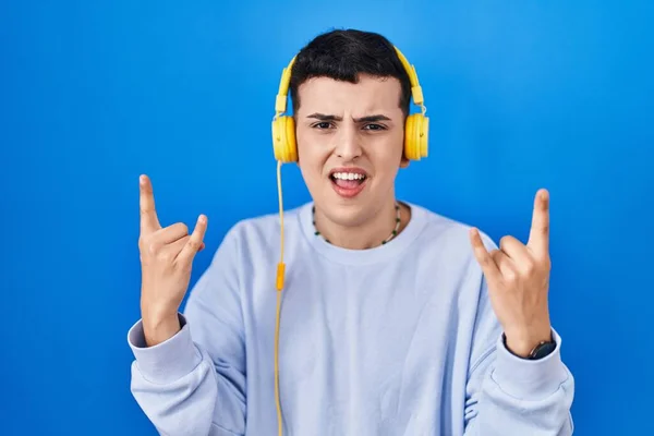 Non binary person listening to music using headphones shouting with crazy expression doing rock symbol with hands up. music star. heavy music concept.