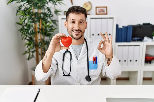 Młody Lekarz Trzymający Serce Klinice Medycznej Robi Znak Palcami Uśmiechając — Zdjęcie stockowe