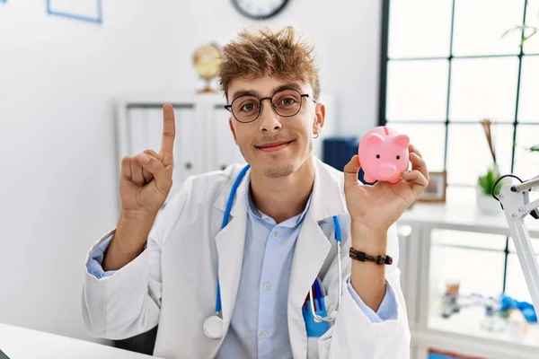 Young caucasian doctor man wearing doctor uniform holding piggy bank at the clinic smiling with an idea or question pointing finger with happy face, number one