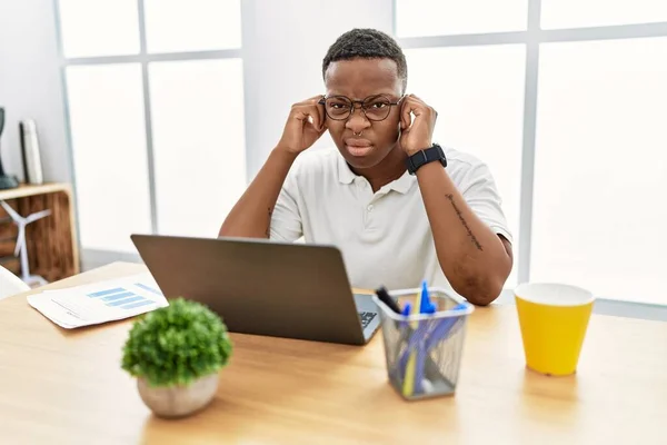Young african man working at the office using computer laptop covering ears with fingers with annoyed expression for the noise of loud music. deaf concept.
