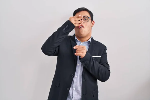 Young hispanic man with down syndrome wearing business style peeking in shock covering face and eyes with hand, looking through fingers with embarrassed expression.