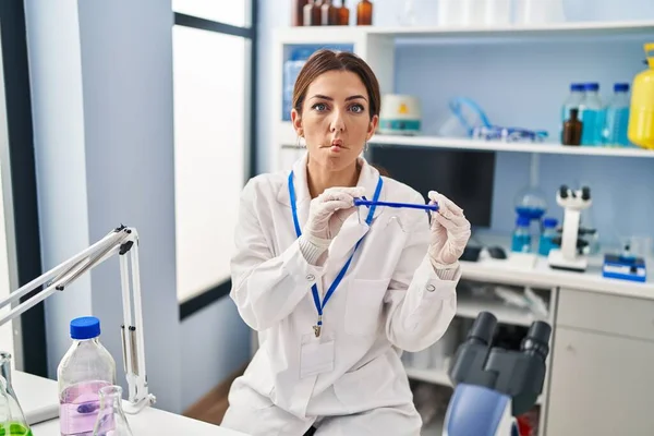 Young brunette woman working at scientist laboratory wearing safety glasses making fish face with mouth and squinting eyes, crazy and comical.
