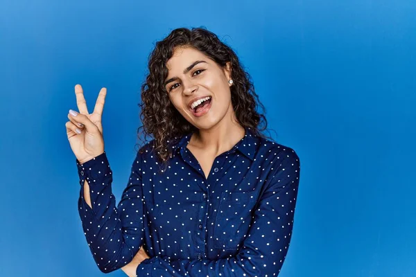 Young brunette woman with curly hair wearing casual clothes over blue background smiling with happy face winking at the camera doing victory sign. number two.