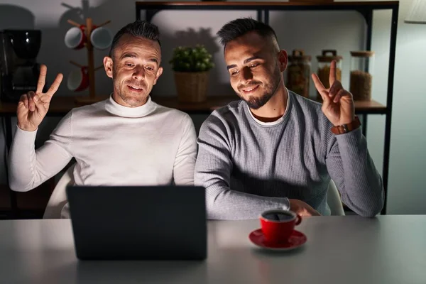 Homosexual couple using computer laptop smiling looking to the camera showing fingers doing victory sign. number two.