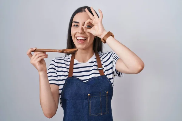 若いですブルネット女性身に着けているエプロンテイスティング食品保持木製スプーン笑顔幸せな行いますOk Signとともに手で目を通して指を見る — ストック写真