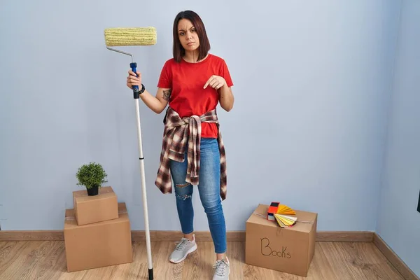 Young hispanic woman painting home walls with paint roller pointing down looking sad and upset, indicating direction with fingers, unhappy and depressed.