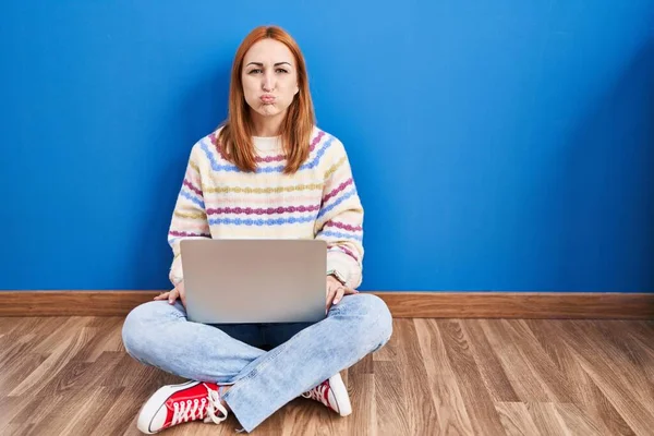 自宅でノートパソコンを使用している若い女性が面白い顔で頬をむき出しの床に座っている 口は空気で膨らみ狂気の表情で — ストック写真