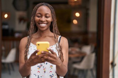 Afro-Amerikalı bir kadın kahve dükkanının terasında akıllı telefon kullanarak gülümsüyor.