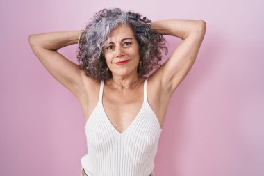 Pembe arka planda duran gri saçlı orta yaşlı kadın rahatlıyor ve esniyor, kolları ve elleri başının arkasında mutlu bir şekilde gülümsüyor. 