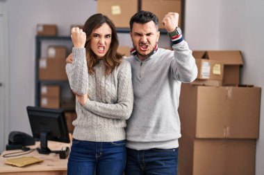 Young hispanic couple working at small business ecommerce annoyed and frustrated shouting with anger, yelling crazy with anger and hand raised 