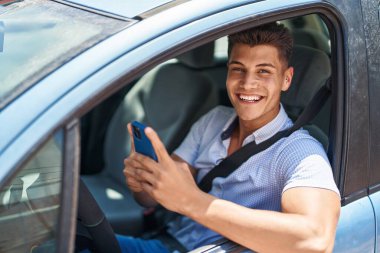 Young hispanic man using smartphone sitting on car at street