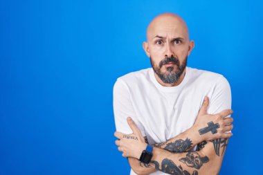 Mavi arka planda dikilen İspanyol dövmeli adam yüzünde üzgün bir ifadeyle kış soğuğundan titriyor ve donuyor. 