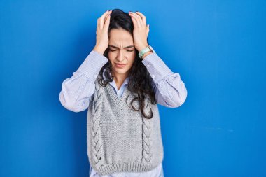 Genç esmer kadın mavi arka planda duruyor. Baş ağrısı, çaresiz ve stresli. Çünkü acı ve migren ağrısı var. Eller başının üstünde.. 