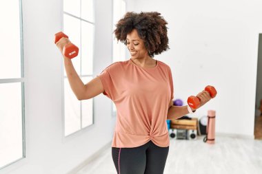 Genç Afrikalı Amerikalı kadın gülümsüyor spor merkezinde halterleri kullanarak kendine güven eğitimi alıyor