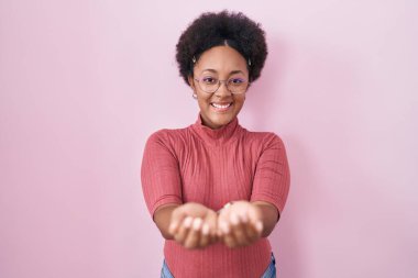 Pembe arka planda duran kıvırcık saçlı güzel Afrikalı kadın avuçlarını birbirine uzatarak gülümsüyor ve jest yapıyor. Bekleme ve koruma 
