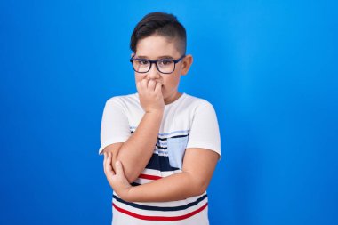 Young hispanic kid standing over blue background looking stressed and nervous with hands on mouth biting nails. anxiety problem. 