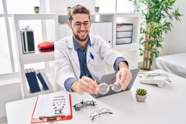 Young man optician holding glasses using laptop at clinic