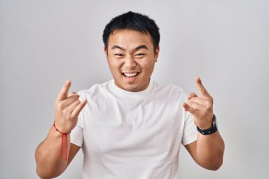 Young chinese man standing over white background shouting with crazy expression doing rock symbol with hands up. music star. heavy music concept. 