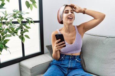 Genç ve güzel bir kadın akıllı telefondan mesaj yazıyor. Kanepede oturuyor, gülümsüyor, saçlarına dokunuyor, el hareketi yapıyor, çekici ve modaya uygun poz veriyor. 