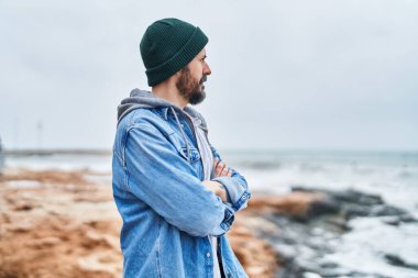 Kel genç adam sahilde kollarını kavuşturmuş el kol hareketi yapıyor.